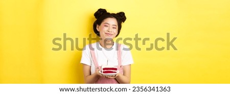 Happy asian birthday girl with bright makeup, blowing candle on cake, making wish, standing on yellow background.