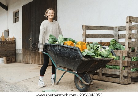 Photo of young woman pushing wagon full pumpkin and cabbage on farm outdoors. Smiling female farm worker holding a wheelbarrow with organic harvest production