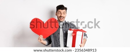 Valentines day and love concept. Handsome man holding gift box and red heart cutout for lover, going on romantic date, smiling excted and looking at empty space, white background. Royalty-Free Stock Photo #2356348913