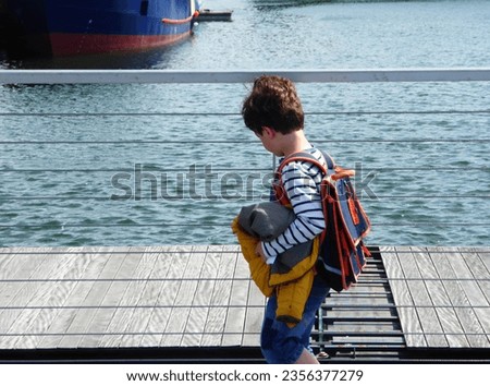 Boy walking and dressed in a sailor top, carrying a yellow coat and a satchel. Sea and boat in the background.