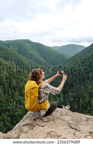 Young traveler with a yellow backpack is resting and taking pictures on the phone on a mountain cliff, admiring the landscape of the high Tatras, mountain range. Beautiful woman on top of the mountain
