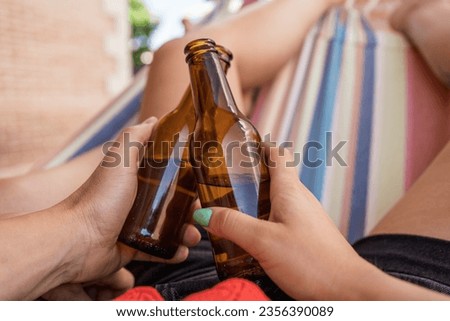 Unrecognizable couple with a beer in their hammock. Close-up point of view image of the hands of a boy and a girl toasting with a few bottles of fresh beers. Royalty-Free Stock Photo #2356390089
