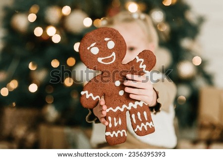 Merry Christmas and Happy Holidays. Child girl eating big iced ingerBread man cookies under Christmas tree Waiting for Christmas. Celebration New Year.