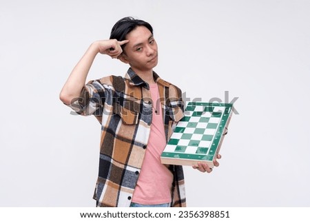 A cocky young asian man pointing to his smarts while holding a professional chessboard. Isolated on a white background.