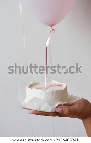 Bento cake with a burning candle in a female hand against the background of pink balls and a white wall. Holiday birthday. Woman holding a birthday cake
