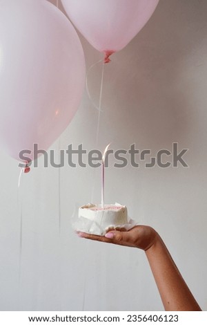 Bento cake with a burning candle in a female hand against the background of pink balls and a white wall. Holiday birthday. Woman holding a birthday cake