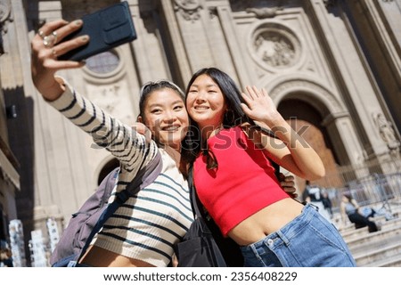 Cheerful young Asian women tourists in casual outfits smiling and taking self portrait on smartphone while standing against Cathedral of Granada in Spain during holidays