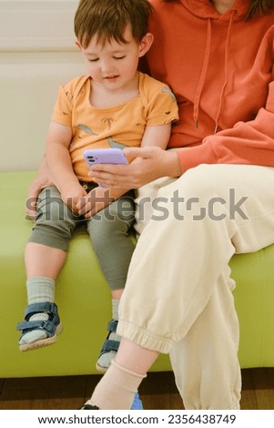 The mother and her baby are in the hospital waiting room, reading a book on the mobile phone together while they wait for their doctor's visit. Kid boy aged two years (two-year-old)