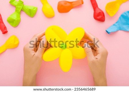 Young adult woman hands holding beautiful flower head created from yellow balloon on light pink table background. Pastel color. Closeup. Point of view shot. Closeup. Making decoration elements.