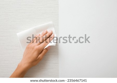 Young adult woman hand holding white dry paper napkin and wiping surface from glue after light gray wallpaper gluing. Closeup. Front view. Repair work of home. Royalty-Free Stock Photo #2356467141