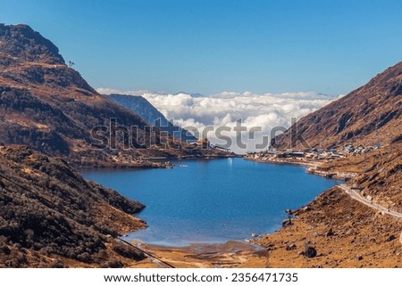 A blue water Tsomgo lake surrounded by desert brown mountains is located high above clouds. The winding road to the distant village leads a way to heaven in cumulus clouds and blue sky. Sikkim, India
