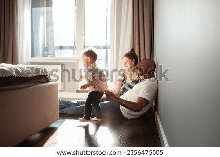 Diverse multiracial family playing with child son at home, authentic family life, unposed, spontaneous moments. Baby learning walk and dad supports him, helps. Royalty-Free Stock Photo #2356475005