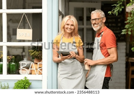 Mature couple small business owners standing at entrance door to their bar with open sign board, happy man and woman wearing aprons smiling and looking at camera, holding coffee and digital tablet