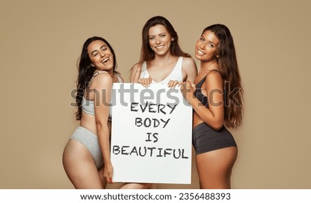 Positive young multiethnic women in different underwear, enjoy natural beauty, hold banner inscription every body is beautiful, isolated on beige background, studio. Lifestyle, health care, love Royalty-Free Stock Photo #2356488393