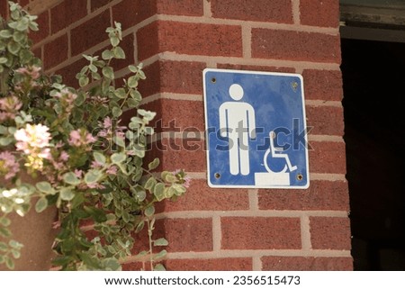 male bathroom washroom mens room square blue white symbol sign and handicap on brick wall outside outdoors exterior next to open bathroom door and branches leaves, close up