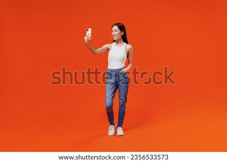Full size young woman of Asian ethnicity 20s in white tank top get video call use mobile cell phone do selfie talk conducting pleasant conversation isolated on plain orange background studio portrait