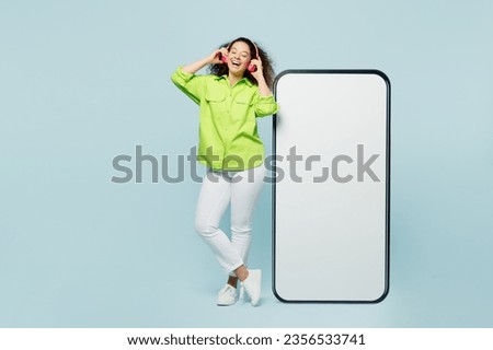 Full body young latin woman wear green shirt casual clothes big huge blank screen mobile cell phone smartphone with workspace area listen to music in headphones isolated on plain blue cyan background