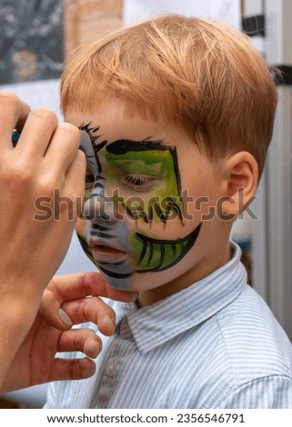 The face of a little boy with a pattern on his face. The artist draws a picture of a kind, green dragon on the face of a child