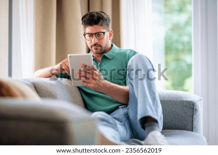 Middle-aged man sitting at home on the sofa holding a digital tablet in his hand. Confident male wearing glasses and casual clothes. He is having video call or browsing on the internet. Full length Royalty-Free Stock Photo #2356562019