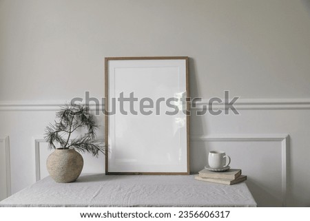 Moody Christmas Scandinavian interior. Minimal winter artistic composition. Blank vertical wooden picture frame mockup, cup of coffee on table. Pine tree branches, old clay vase. White wall background