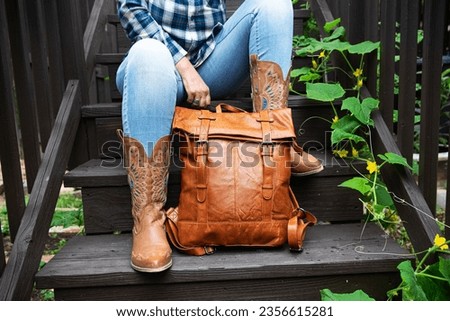 woman in blue jeans and cowboy boots sits on the steps and holds a leather backpack. Close-up. Royalty-Free Stock Photo #2356615281
