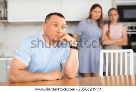 Focused man is sitting in kitchen and prop chin with fist. Father comes up with thinking about options for solving difficult situation of crisis. Family stand behind husband and ask about what Royalty-Free Stock Photo #2356623999