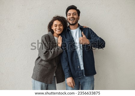 Stylish happy multiracial couple of friends embracing, standing together showing thumb up. Smiling business colleagues looking at camera. Portrait of Indian man and African American woman Royalty-Free Stock Photo #2356633875