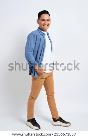Full length of smiling young Asian man in casual shirt walking with hand in pocket and confidently looking at camera isolated on white background