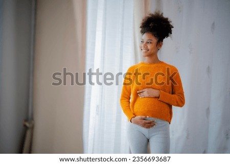 African American pregnancy woman in yellow sweater and gray legging standing beside glasses door, embrassing her belly, looking outside with smile of happiness.