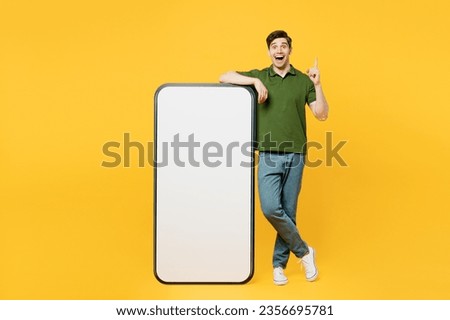 Full body young smart happy man he wears green t-shirt casual clothes big huge blank screen mobile cell phone smartphone with workspace area point index finger up isolated on plain yellow background