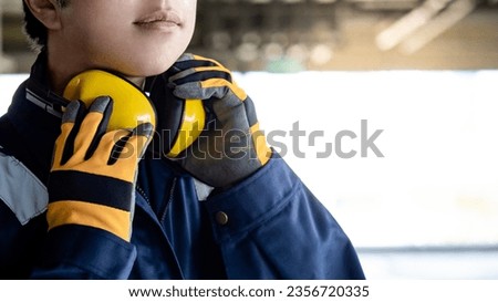 Asian man construction worker wearing uniform suit, safety helmet, goggles and protective gloves holding yellow ear muffs or ear defenders on his neck at construction site Royalty-Free Stock Photo #2356720335