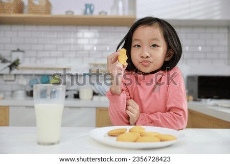 Kid asian black hair girl sitting on table and drinking milk while eating vanilla cookies for breakfast with enjoying time. Tasty food and delicious food with happy meal lifestyle kid concept