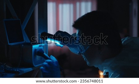 A male expert criminologist removes fingerprints from a jewelry box with a brush. A man gathers evidence using an ultraviolet lamp at a crime scene in a dark apartment lit by red, blue police sirens. Royalty-Free Stock Photo #2356748011