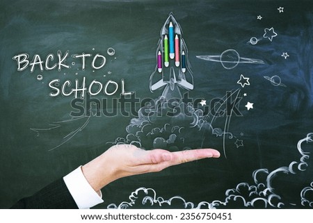 Close up of male hand holding abstract back to school sketch with pencils and rocket on chalkboard wall background. Education and knowledge concept