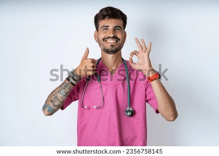 young caucasian doctor man wearing pink medical uniform feeling happy, amazed, satisfied and surprised, showing okay and thumbs up gestures, smiling