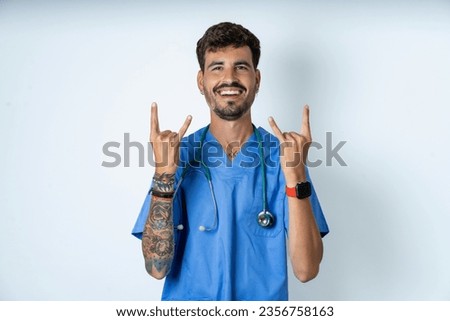 young caucasian man wearing blue medical uniform makes rock n roll sign looks self confident and cheerful enjoys cool music at party. Body language concept. Royalty-Free Stock Photo #2356758163