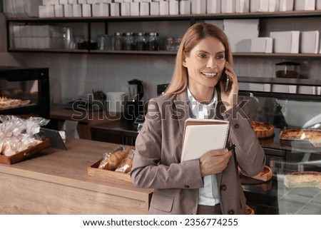 Happy business owner with notebook and pen talking on phone in bakery shop