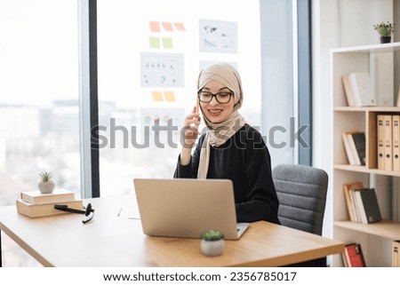 Joyful lady in traditional muslim headscarf speaking on smartphone while reading report from digital screen. Experienced employee coming up with idea of improving company productivity in workplace.