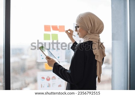 Side view of arabian office administrator with tablet writing brief notes on stickers attached to window glass. Stylish entrepreneur in hijab analysing accounting records via infographics.