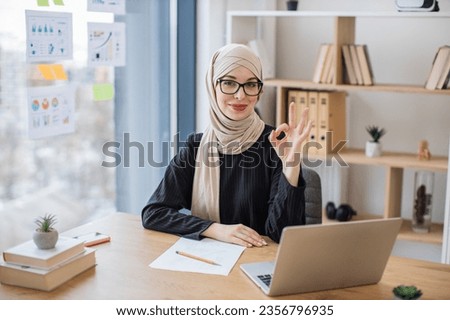 Portrait of attractive arabian woman in glasses posing behind wireless gadget during working process, showing sign ok