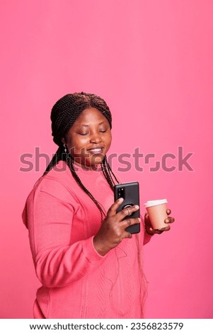 Smiling woman with pink sweather holding cup of coffee while reading the news on phone, posting picture social media. Young adult watching funny video on smartphone whle posing in studio