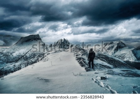 Adventure of mountaineer standing on top of snowy mountain with dark stormy sky in winter on Segla mount at Senja Island, Norway Royalty-Free Stock Photo #2356828943