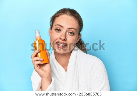 Young blonde woman in a robe holding a sunscreen in a studio Royalty-Free Stock Photo #2356835785