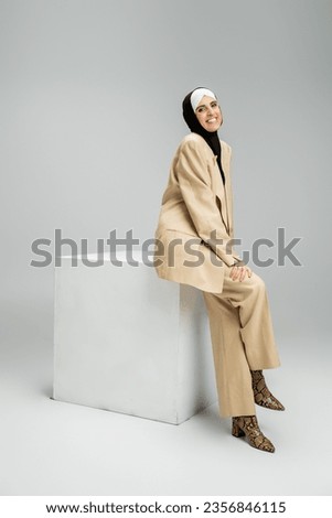 joyful muslim businesswoman in hijab and beige suit sitting on white cube on grey, full length