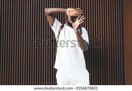 Black man in white shirt and white shorts making frame hands on air while standing against brown metal wall in daylight outdoors