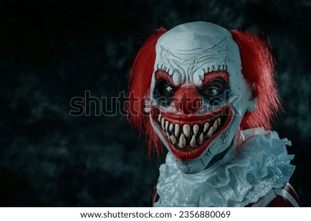 a mad evil redhead clown, wearing a white and red striped costume with a white ruff, stares at the observer with a creepy smile, in front of a dark background Royalty-Free Stock Photo #2356880069