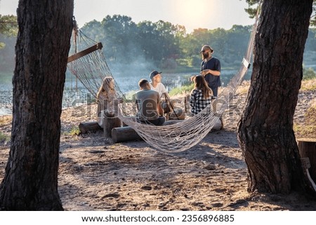 A stretched hammock on the trees against the background of a company of friends vacationing in nature in the summer on the banks of the river, campfire