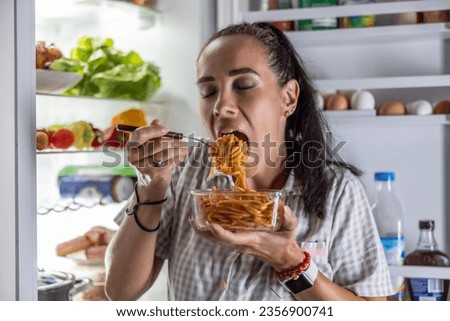 A very hungry woman in pajamas is enjoying spaghetti at the refrigerator at night. Royalty-Free Stock Photo #2356900741