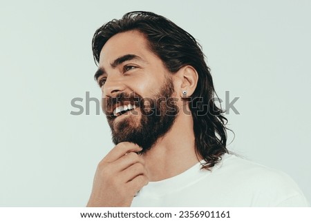 Man with a beard and a radiant skin tone stands in a studio, wearing a perfect smile on his face. Happy young man embracing his beauty and masculine looks, expressing confidence and self-assurance. Royalty-Free Stock Photo #2356901161