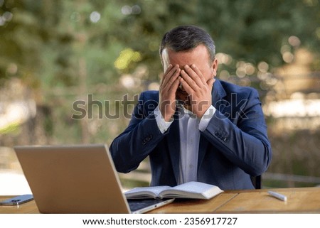 a mature business man in an outdoor ambition working in a laptop and phone ,blue coat suit,multiracial asian, praying with two hands in his face ,less freespace .